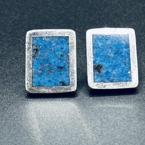 Puede incluir: Un par de pendientes rectangulares con un marco de metal plateado que rodea una incrustación de piedra azul vibrante y texturizada con sutiles motas negras. El diseño es simétrico, con cada pendiente mostrando un borde metálico pulido y una pieza central rectangular plana de material mineral azul.
