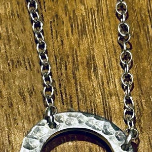 May include: A silver-toned necklace featuring a circular pendant with a hammered, textured surface. The pendant is suspended from a delicate chain and includes three small, smooth silver-toned rings attached to the bottom of the main circle.