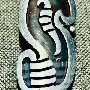 Pode incluir: Um broche de metal prateado com o formato de uma letra S estilizada. O design incorpora um elemento curvo em relevo que se assemelha a um polegar ou gancho na parte inferior e uma forma pontiaguda e afilada no topo sobre um fundo escuro.