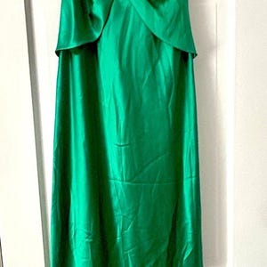 Könnte beinhalten: Ein bodenlanges, ärmelloses smaragdgrünes Kleid mit V-Ausschnitt und drapiertem Oberteil. Die Taille ist mit einer silberfarbenen Zierbrosche mit klaren Kristallen versehen. Der Rock ist lang und gerade und aus glattem, satinartigem Stoff.