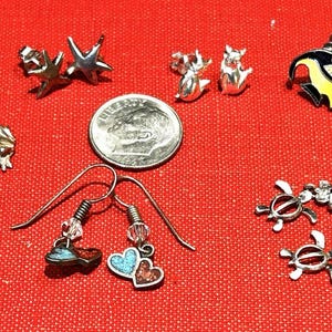 May include: A collection of silver-toned earrings including frog dangles, star studs, small animal studs, black and yellow fish studs, heart dangles with blue and red inlay, and turtle dangles with clear crystals, shown with a US dime for scale.