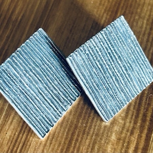 May include: A pair of square-shaped silver-toned earrings featuring a textured surface with vertical, raised parallel lines. The earrings have a metallic finish and a geometric design, presenting a clean, sharp edge on all four sides of each piece.