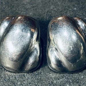 Può includere: Un paio di orecchini argentati e strutturati. Ogni orecchino ha una forma oblunga arrotondata con un design attorcigliato e scanalato. Gli orecchini sono esposti su una superficie scura e strutturata.