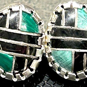 May include: A pair of oval-shaped earrings with a silver-colored metal frame. The earrings feature a mosaic design with black and green stone inlays. The green stones have a marbled appearance. The earrings are displayed on a black surface.