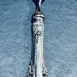 May include: A silver fork with an ornate handle. The handle features detailed floral and scrollwork designs. The fork tines are long and slender. The silverware is likely vintage or antique.