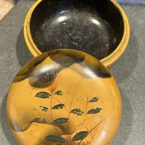 May include: A round, lidded container with a yellow exterior and a black interior. The lid features a painted landscape with stylized plants and foliage in shades of green, brown, and yellow. The container is open, revealing its dark interior.