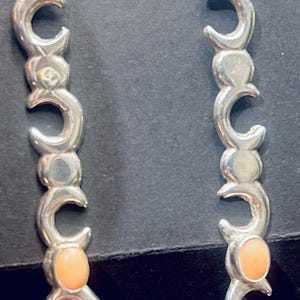 May include: Silver-toned dangle earrings featuring a series of crescent moon shapes connected vertically. Each earring has a small, oval, peach-colored stone at the bottom. The earrings hang from a simple hook.