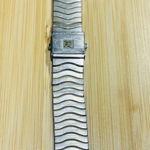 Könnte beinhalten: Ein silberfarbenes Metall-Uhrenarmband mit einem sich wiederholenden wellenförmigen, texturierten Muster über die Glieder. Das Armband verfügt über einen zentralen rechteckigen Verschlussmechanismus mit einem kleinen, geprägten goldenen Logo.