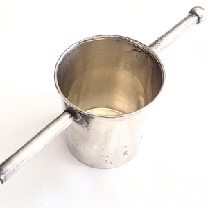 Könnte beinhalten: Ein silberfarbener Cocktail-Jigger mit einem zylindrischen Becher und einem Griff, der sich auf beiden Seiten erstreckt. Jedes Griffende hat eine kleine, runde Kugel. Der Jigger wird wahrscheinlich zum Abmessen von Zutaten verwendet.