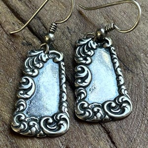 May include: A pair of silver-toned dangle earrings featuring a rectangular pendant with rounded corners and an ornate, raised scrollwork border. The center of each pendant is smooth and recessed, and the earrings hang from simple silver-toned hooks.