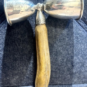 Könnte beinhalten: Ein Vintage-Cocktail-Jigger aus Silber mit einem natürlichen Geweihgriff. Der Jigger hat zwei verspiegelte Messbecher und ein dekoratives Silberband am Griffansatz. Der Griff ist hellbraun.