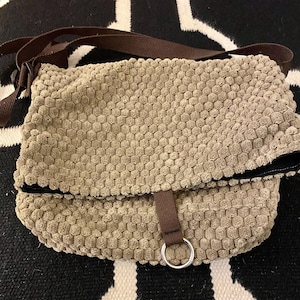 May include: A beige crossbody bag featuring a textured, honeycomb-like fabric surface. The bag includes a brown fabric strap, a fold-over flap, and a brown fabric tab closure secured with a silver-toned metal ring at the bottom center.
