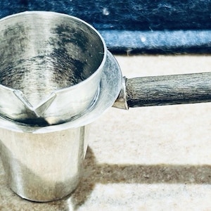 Könnte beinhalten: Ein silberner Kaffeeausgießer mit Holzgriff. Der Ausgießer hat einen kleinen Ausguss und einen kreisförmigen Boden. Der Griff ist aus dunklem Holz. Der Ausgießer steht auf einer hellen Oberfläche.