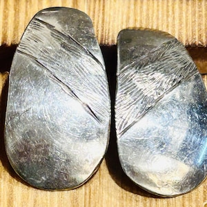 Pode incluir: Um par de brincos prateados de formato abstrato. Cada brinco tem um acabamento em metal escovado texturizado com detalhes gravados diagonais e lineares. Os brincos são exibidos em uma superfície de madeira.
