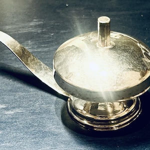 Könnte beinhalten: Eine polierte, silberfarbene Metallglocke mit einer abgerundeten Kuppel, einem kleinen zylindrischen Griff in der Mitte der Kuppel und einem langen, flachen, gebogenen Hebel an der Seite, mit reflektierender Oberfläche und rundem Sockel.