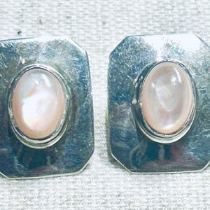 May include: A pair of rectangular silver earrings featuring a polished metallic finish and a central oval-shaped iridescent mother-of-pearl cabochon stone set within a raised bezel. The earrings have a smooth, reflective surface and rounded corners.