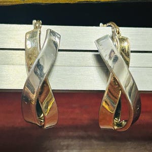 May include: A pair of hoop earrings featuring a polished silver-tone metal finish. The design consists of two intertwined, ribbon-like bands that cross over each other to form a continuous, fluid loop shape with a hinged closure at the top.