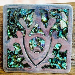Op de afbeelding: Vierkante zilverkleurige hanger met een abalone schelpinlegsel. De hanger heeft een uitgesneden ontwerp van een gestileerde figuur. De abaloneschelp toont iriserende blauwe, groene en paarse tinten, wat een opvallend contrast met het zilver creëert.
