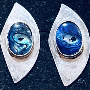 May include: A pair of silver earrings in a teardrop design. Each earring has an oval, iridescent blue abalone shell centre, set within a gold-coloured metal and a silver border. The earrings are shown against a dark background.