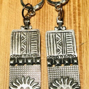 May include: A pair of silver-toned metal earrings featuring rectangular pendants with a textured surface. The design includes vertical stripes, a row of small studs, a grid pattern, and a stylized sun face at the bottom, attached to circular hoops.