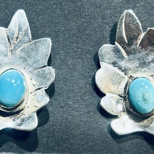 May include: A pair of silver earrings shaped like stylized leaves, each featuring a central oval-shaped turquoise stone set in a smooth bezel. The silver metal has a polished finish with etched lines detailing the leaf veins and edges.