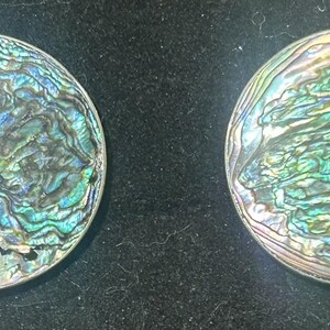 May include: Two circular abalone shell discs featuring a natural iridescent finish with swirling patterns of blue, green, purple, and silver. The surfaces display a high-gloss, multi-colored luster with organic textures and unique color variations.