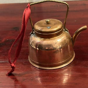 Op de afbeelding: Een miniatuur koperkleurig metalen ketelornament met een ronde behuizing, een kleine tuit en een afneembaar deksel met een centrale knop. Een gestructureerd goudkleurig metalen handvat is aan de bovenkant bevestigd, met een dun rood lint aan één kant.