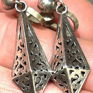 May include: A pair of silver-toned drop earrings with a geometric design. Each earring has a diamond-shaped pendant with detailed openwork filigree patterns. The earrings have a ball-shaped stud and a clip-on mechanism.