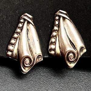 May include: A pair of silver-toned earrings. Each earring has a stylized, asymmetrical shape with a row of small, round beads along one edge. The design includes a swirl detail at the bottom and a textured top.