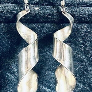May include: A pair of silver dangle earrings featuring a spiraling, ribbon-like design. Each earring hangs from a thin silver hook and displays a polished, metallic finish with a visible stamp on the inner surface of the left earring.