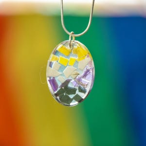 May include: An oval-shaped pendant suspended from a silver-toned chain. The pendant features a clear resin base embedded with small, irregular fragments of yellow, white, purple, and dark green material, creating a mosaic-like appearance.