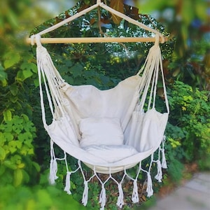 May include: A cream-coloured hammock chair with a wooden bar and white rope supports. The chair has a soft, cushioned seat and a matching pillow. Tassels hang from the bottom edge. The background is lush green foliage.