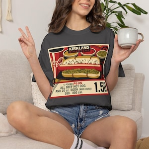 May include: Dark gray t-shirt featuring a vintage-style advertisement for a Kirkland Signature hot dog combo. The graphic includes a hot dog, soda, and the price of $1.50. The text reads "¼ POUND PLUS ALL BEEF HOT DOG AND 20 oz. SODA (With Refill) 580 - 850 cal."