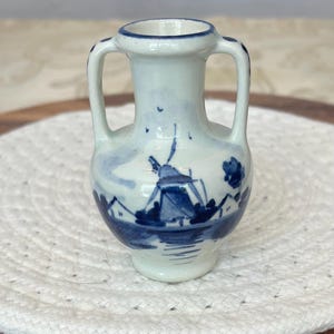 May include: A small white ceramic vase featuring two handles and a blue hand-painted design of a traditional windmill landscape. The vessel has a narrow neck, a rounded body, and a blue rim detail, showcasing a classic delft-style aesthetic.