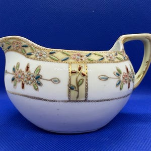 May include: A white porcelain cream jug featuring a gold-trimmed handle and rim. The body is decorated with a band of raised enamel floral patterns in shades of pink, green, and blue, accented by gold geometric borders and vertical panels.