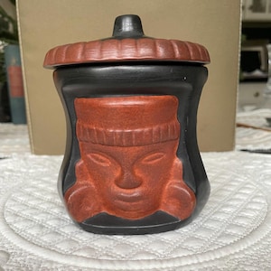 May include: A black ceramic jar with a lid. The jar features a raised, reddish-brown face design on the front. The lid and face design are reddish-brown, while the rest of the jar is black. The jar is sitting on a white, patterned surface.