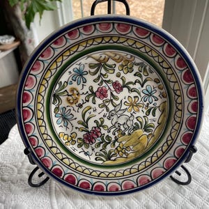 May include: A decorative ceramic plate with a hand-painted floral and rabbit design. The plate features a blue rim, a green and yellow border, and a white center with colorful flowers and a rabbit. The plate is displayed on a black metal stand.