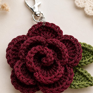 Peut inclure: Un porte-cl&eacute;s en forme de fleur crochet&eacute;e bordeaux fonc&eacute; avec des p&eacute;tales superpos&eacute;s et deux feuilles crochet&eacute;es vertes sur le c&ocirc;t&eacute;. La fleur est reli&eacute;e &agrave; un fermoir mousqueton en m&eacute;tal argent&eacute; sur le dessus pour une fixation facile.