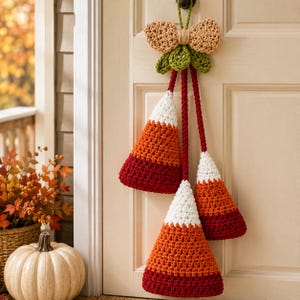 May include: A crochet wall hanging featuring three cone-shaped candy corn ornaments in white, orange, and dark red. The ornaments are suspended by long red cords from a central tan bow and green leaves, all crafted from yarn and hung on a door.
