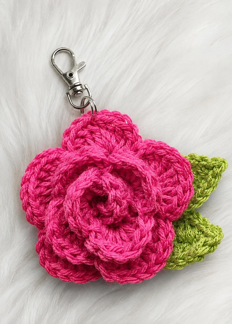 Peut inclure: Un porte-cl&eacute;s fleur au crochet compos&eacute; d'une rose rose vif aux p&eacute;tales superpos&eacute;s et de deux feuilles vert citron textur&eacute;es sur le c&ocirc;t&eacute;. Le motif floral est fix&eacute; &agrave; un mousqueton pivotant en m&eacute;tal de couleur argent&eacute;e.