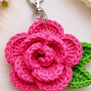 Peut inclure: Un porte-cl&eacute;s fleur au crochet rose vif avec des p&eacute;tales superpos&eacute;s et deux feuilles vertes au crochet, fix&eacute; &agrave; un mousqueton en m&eacute;tal argent&eacute;. La fleur est r&eacute;alis&eacute;e en fil &eacute;pais et textur&eacute;, formant une structure circulaire en relief.