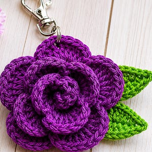 Peut inclure: Un porte-cl&eacute;s fleur crochet&eacute; violet pr&eacute;sentant un motif de rose &agrave; plusieurs couches avec deux feuilles crochet&eacute;es vert vif attach&eacute;es sur le c&ocirc;t&eacute;. La fleur est reli&eacute;e &agrave; un mousqueton en m&eacute;tal argent&eacute; par un petit anneau m&eacute;tallique.