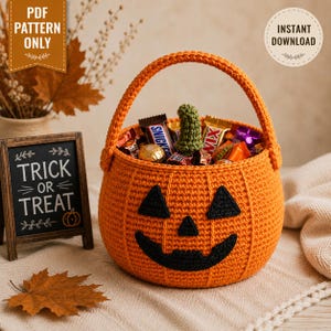 Può includere: Un cestino a forma di zucca lavorato all'uncinetto in arancione, con un volto di jack-o'-lantern nero e un picciolo verde, riempito di barrette di cioccolato. Accanto c'è una piccola lavagna con la scritta TRICK OR TREAT e un disegno.