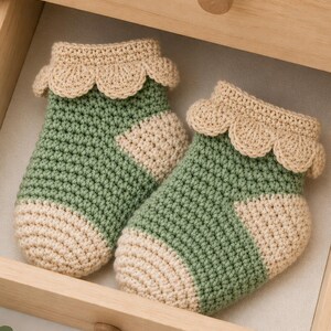 Peut inclure: Une paire de chaussettes de bébé crochetées à la main dans un tiroir. Les chaussettes sont vertes et beiges avec un bord festonné. Les chaussettes sont en fil doux et sont probablement pour un nouveau-né.