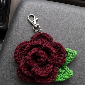 Peut inclure: Une rose au crochet de couleur bordeaux fonc&eacute; avec des p&eacute;tales superpos&eacute;s et deux feuilles au crochet vert vif attach&eacute;es &agrave; la base. La fleur est reli&eacute;e &agrave; un fermoir mousqueton en m&eacute;tal argent&eacute; par un petit anneau m&eacute;tallique circulaire.