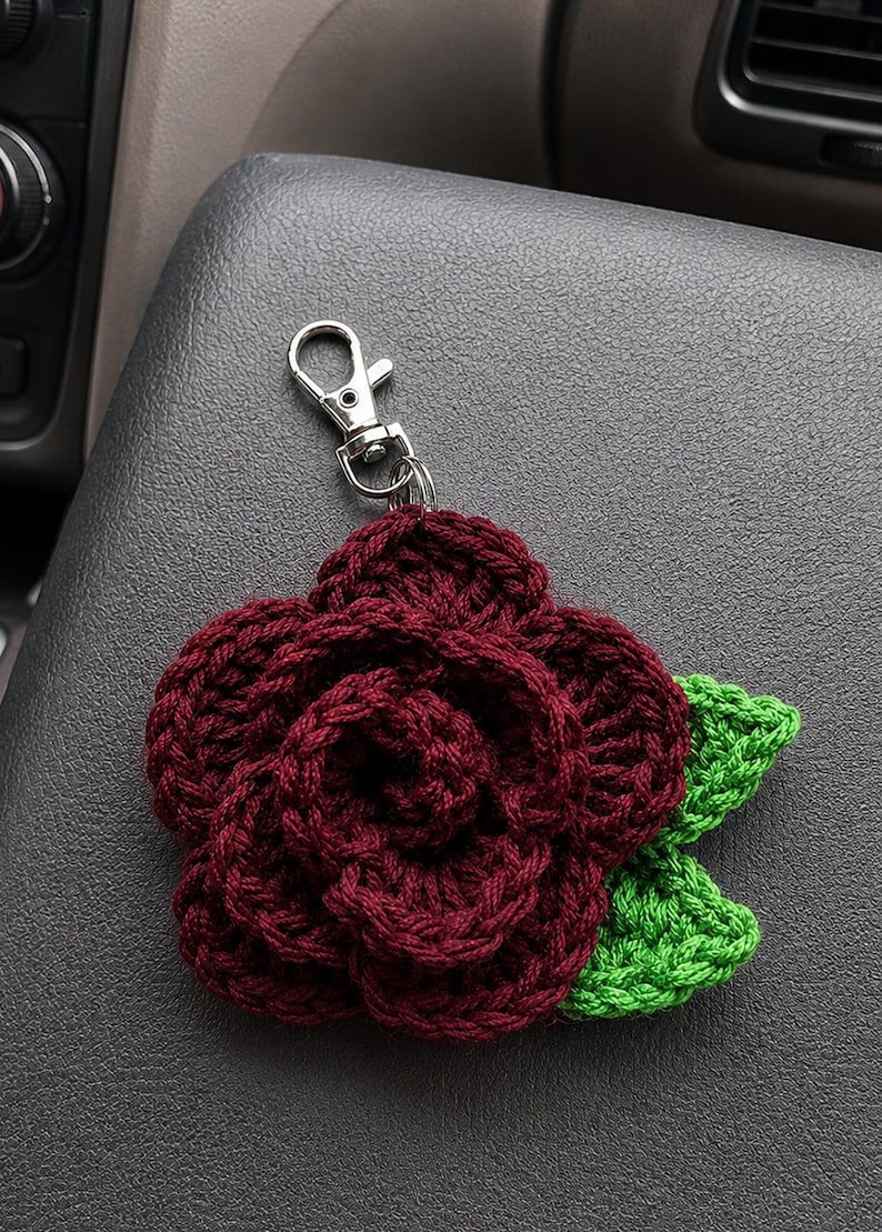 Peut inclure: Une rose au crochet de couleur bordeaux fonc&eacute; avec des p&eacute;tales superpos&eacute;s et deux feuilles au crochet vert vif attach&eacute;es &agrave; la base. La fleur est reli&eacute;e &agrave; un fermoir mousqueton en m&eacute;tal argent&eacute; par un petit anneau m&eacute;tallique circulaire.