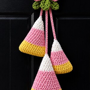 Pode incluir: Uma decora&ccedil;&atilde;o de croch&ecirc; suspensa com tr&ecirc;s pe&ccedil;as em forma de cone nas cores branca, rosa e amarela, penduradas por um la&ccedil;o rosa central e um grupo de folhas verdes. Os tr&ecirc;s cones est&atilde;o dispostos em alturas vari&aacute;veis com padr&atilde;o uniforme.