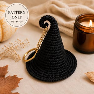Pode incluir: Um porta-chaves de chapéu de bruxa em croché preto com uma ponta enrolada e um anel de metal dourado preso por uma corrente curta. Uma etiqueta branca circular à esquerda apresenta o texto PATTERN ONLY com arte linear e um coração.