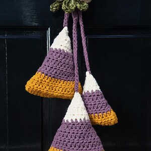Pode incluir: Uma decora&ccedil;&atilde;o de croch&ecirc; suspensa com tr&ecirc;s ornamentos triangulares em forma de doce nas cores branco, roxo e amarelo mostarda. Os ornamentos est&atilde;o suspensos por cord&otilde;es tran&ccedil;ados roxos a um la&ccedil;o de croch&ecirc; bege com detalhes de folhas verdes.