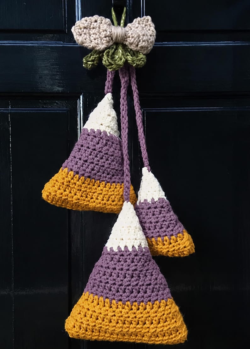 Pode incluir: Uma decora&ccedil;&atilde;o de croch&ecirc; suspensa com tr&ecirc;s ornamentos triangulares em forma de doce nas cores branco, roxo e amarelo mostarda. Os ornamentos est&atilde;o suspensos por cord&otilde;es tran&ccedil;ados roxos a um la&ccedil;o de croch&ecirc; bege com detalhes de folhas verdes.